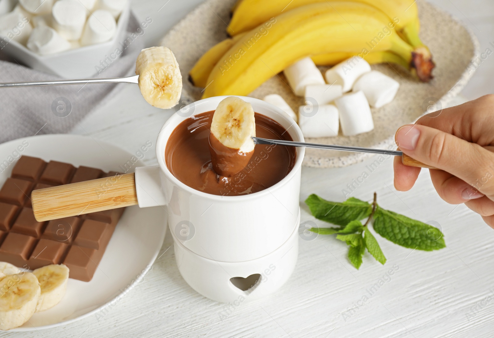 Photo of Woman dipping banana into fondue pot with chocolate at white wooden table, closeup