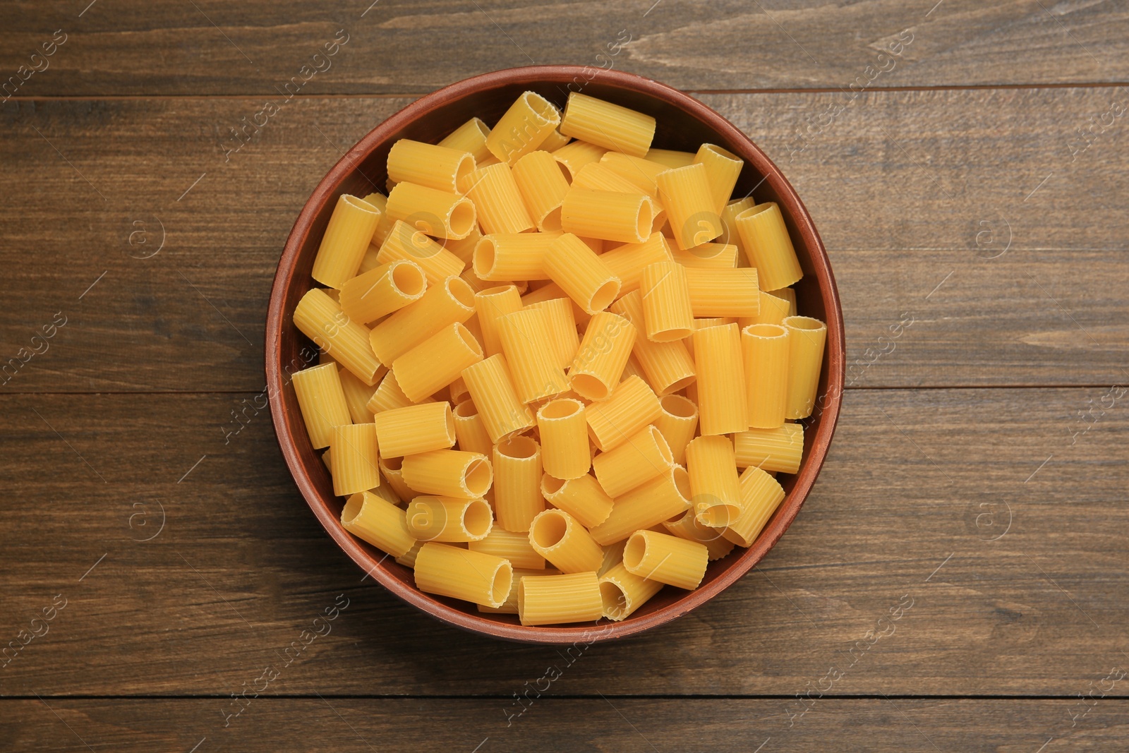 Photo of Raw rigatoni pasta in bowl on wooden table, top view