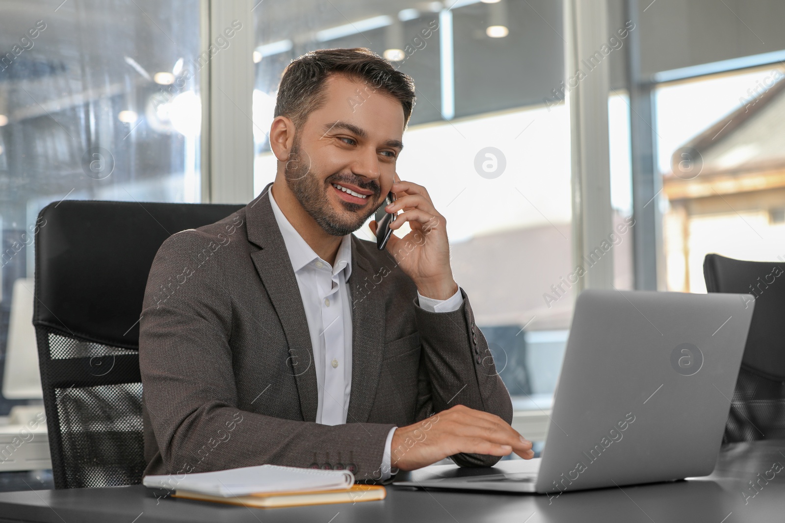 Photo of Happy man using modern laptop while talking on smartphone at black desk in office