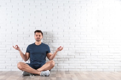 Handsome young man sitting on floor and practicing zen yoga near brick wall, space for text