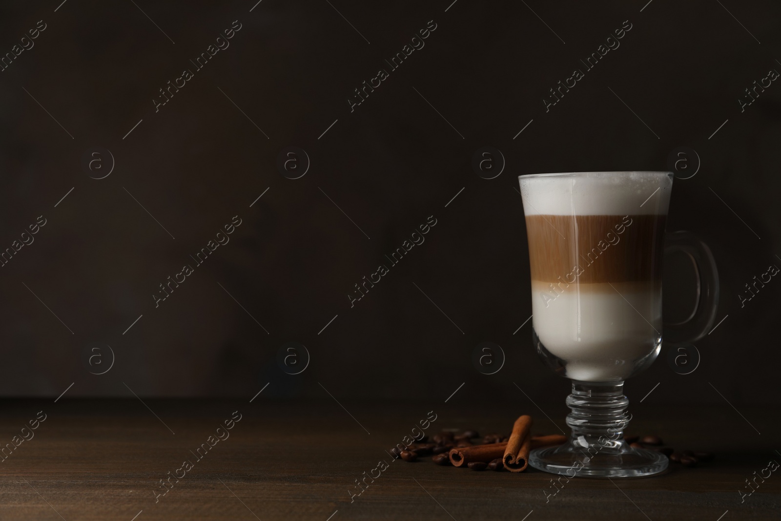 Photo of Hot coffee with milk in glass cup, beans and cinnamon sticks on wooden table. Space for text