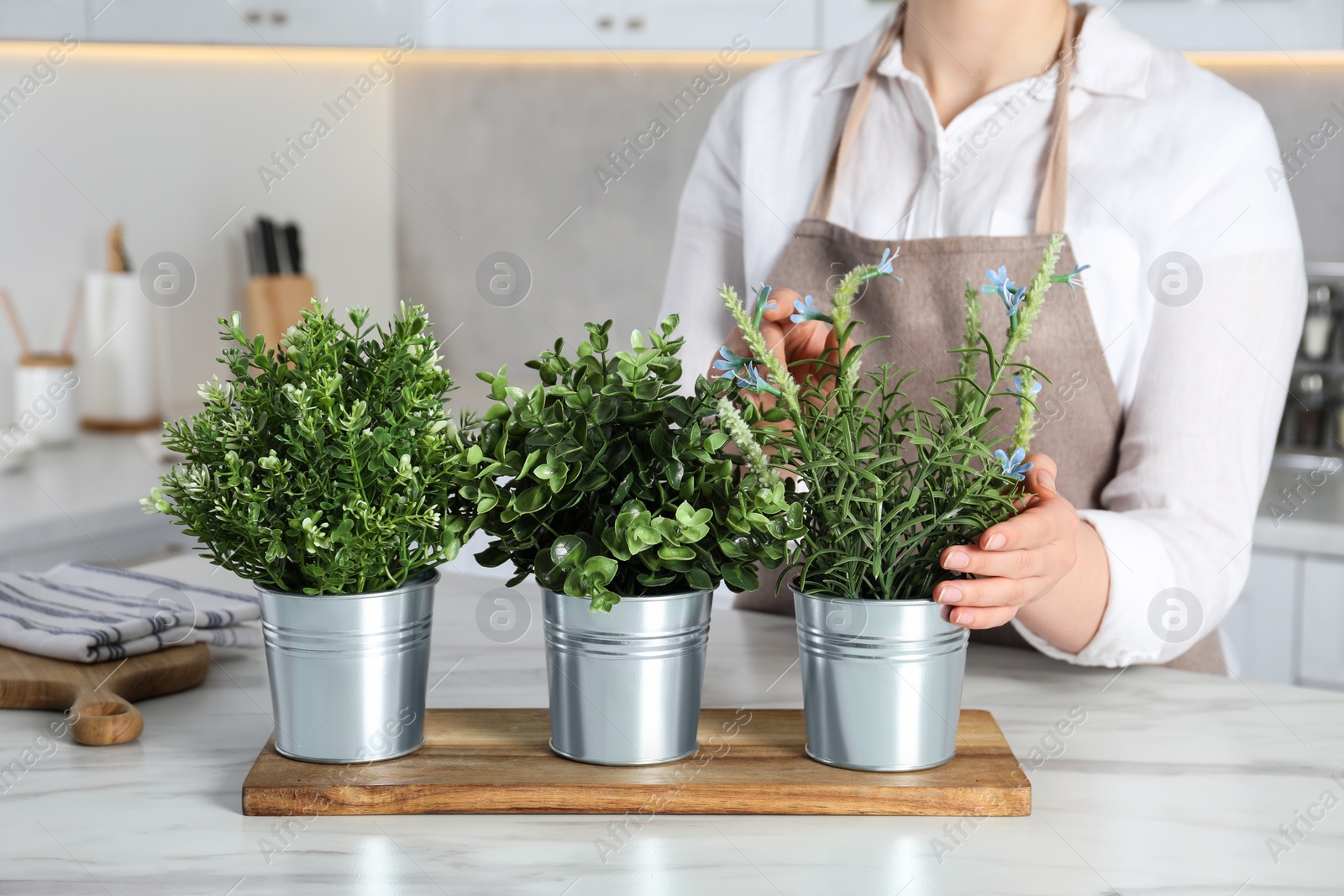 Photo of Woman near white marble table with different artificial potted herbs in kitchen, closeup