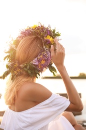 Young woman wearing wreath made of beautiful flowers near river on sunny day