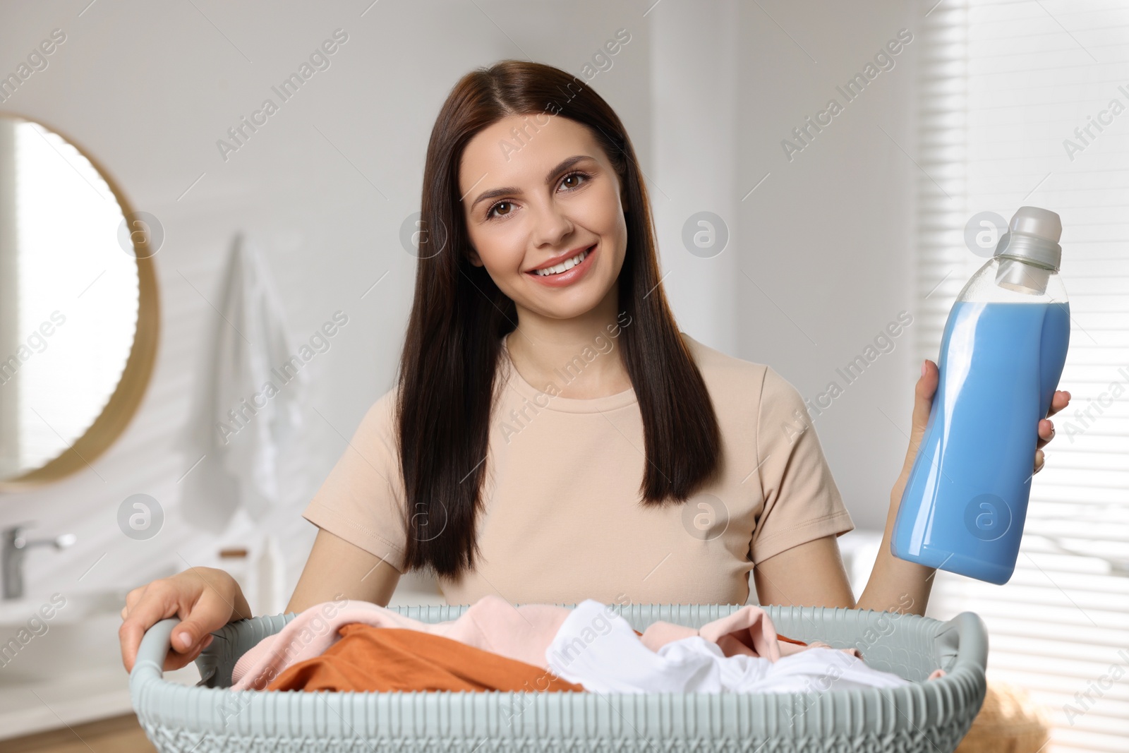 Photo of Woman holding fabric softener and basket with dirty clothes in bathroom