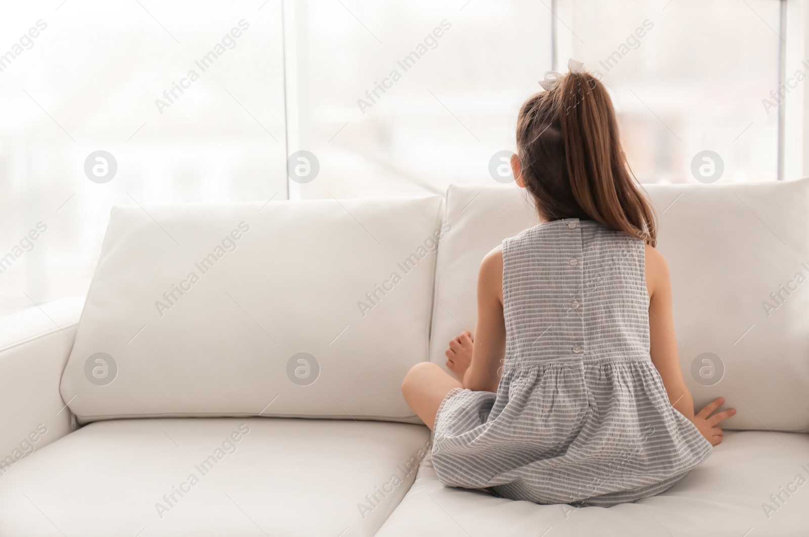 Photo of Lonely little girl sitting on sofa near window indoors. Child autism