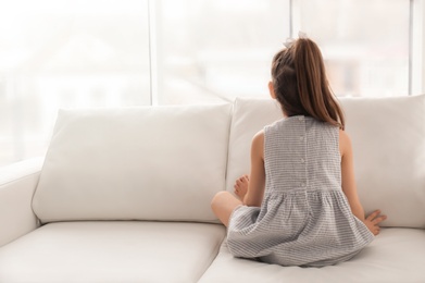 Photo of Lonely little girl sitting on sofa near window indoors. Child autism