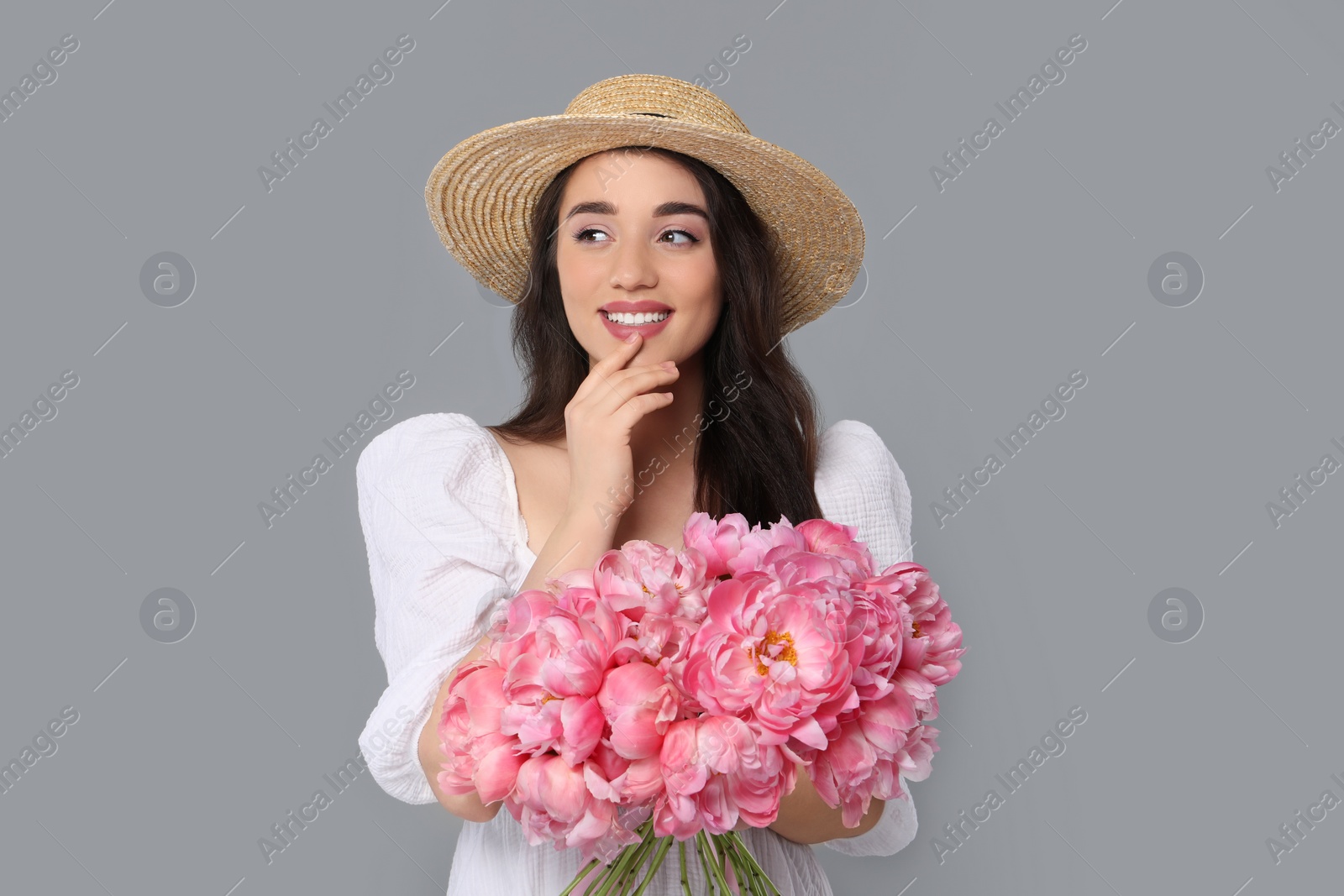 Photo of Beautiful young woman in straw hat with bouquet of pink peonies against grey background