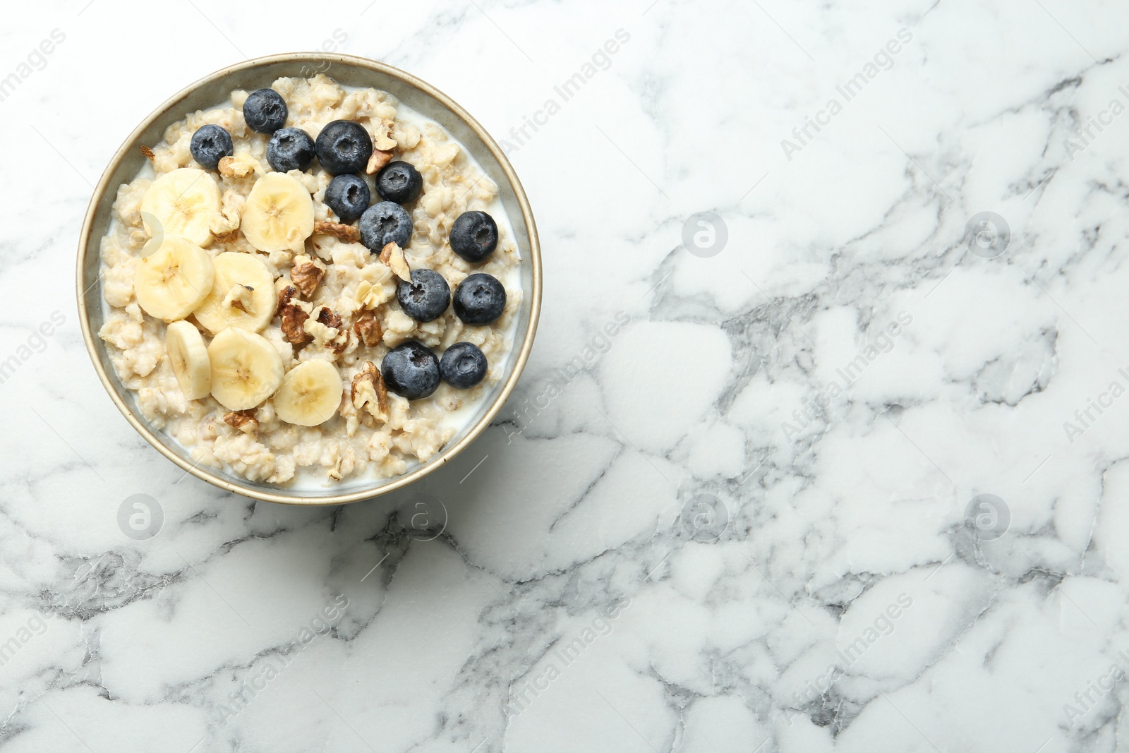Photo of Tasty oatmeal with banana, blueberries, walnuts and milk served in bowl on white marble table, top view. Space for text