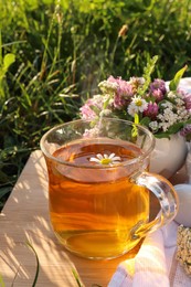 Cup of aromatic herbal tea and ceramic mortar with different wildflowers on wooden board in meadow