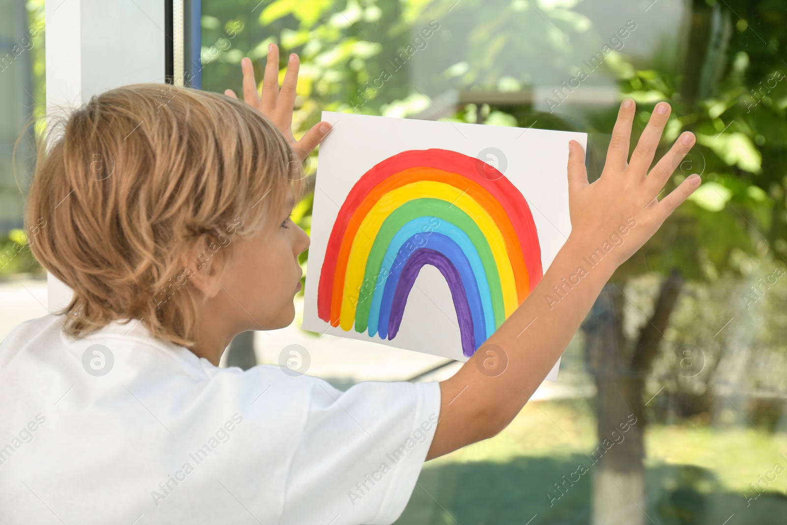 Photo of Little boy holding rainbow painting near window. Stay at home concept