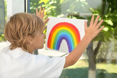 Little boy holding rainbow painting near window. Stay at home concept