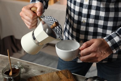 Man pouring aromatic coffee from moka pot into cup at table in kitchen, closeup