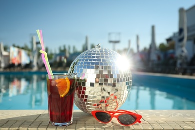 Photo of Shiny disco ball, refreshing cocktail and sunglasses on edge of swimming pool. Party items