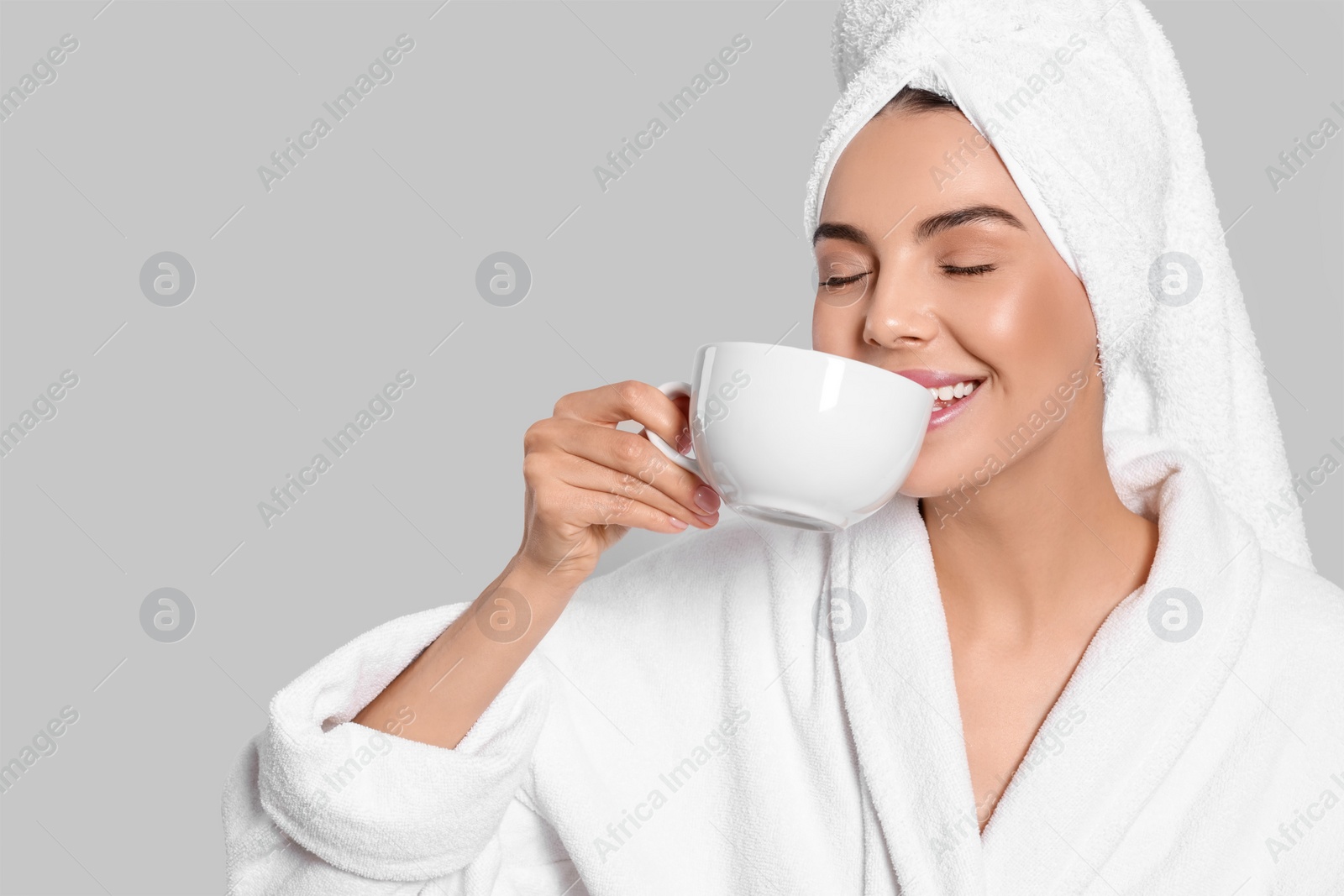Photo of Young woman in bathrobe with towel and cup of drink on light grey background, space for text. Spa treatment