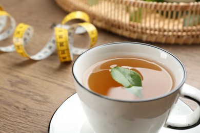 Cup of herbal diet tea and measuring tape on wooden table, closeup