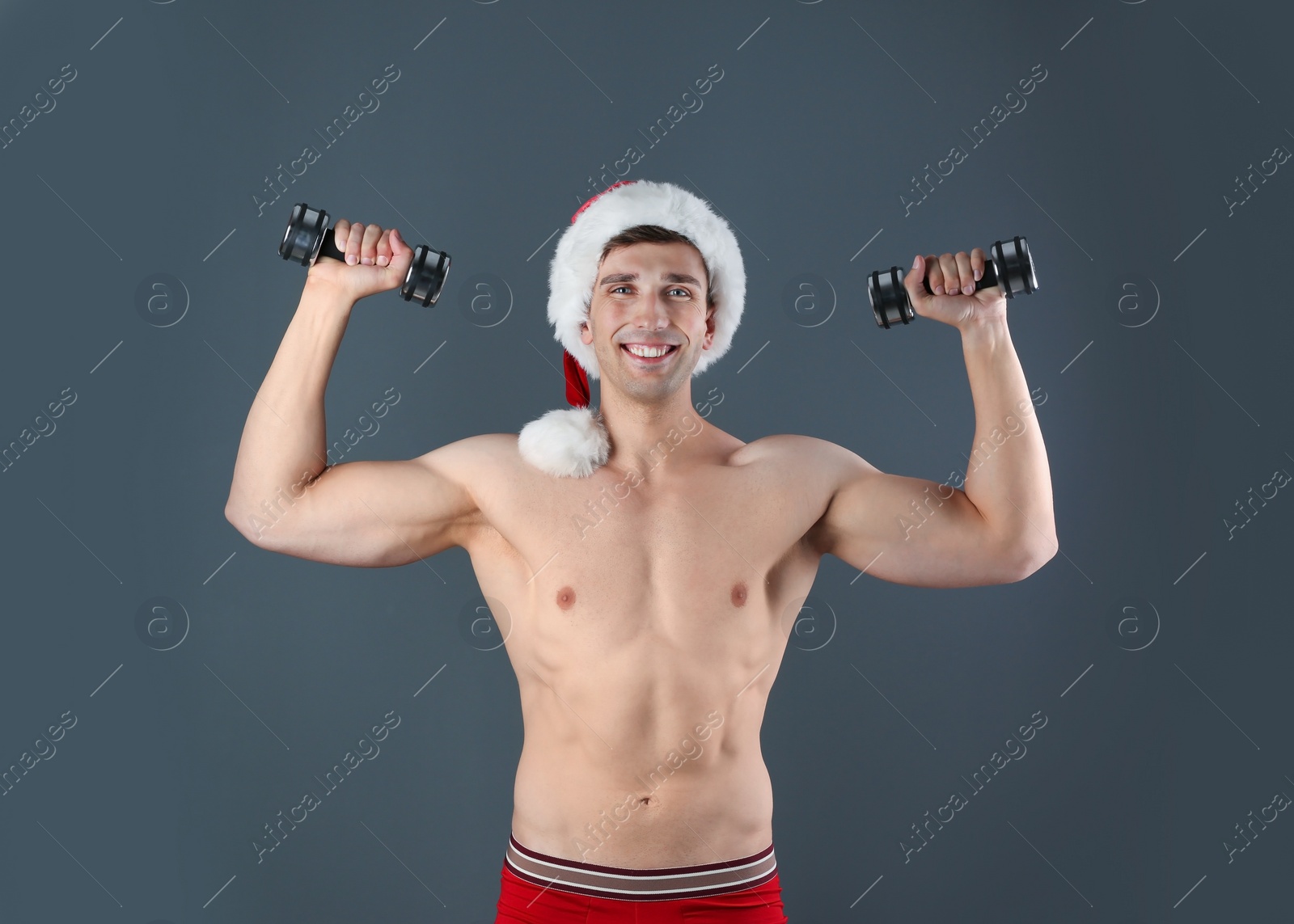 Photo of Young muscular man in Santa hat with dumbbells on gray background