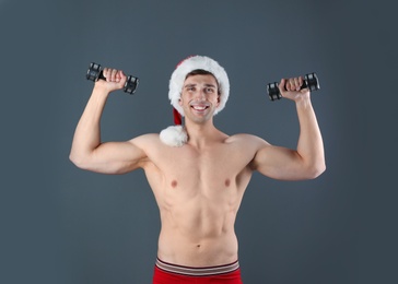 Photo of Young muscular man in Santa hat with dumbbells on gray background