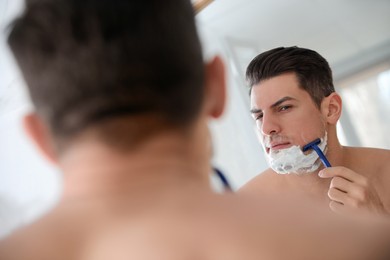 Handsome man shaving near mirror in bathroom