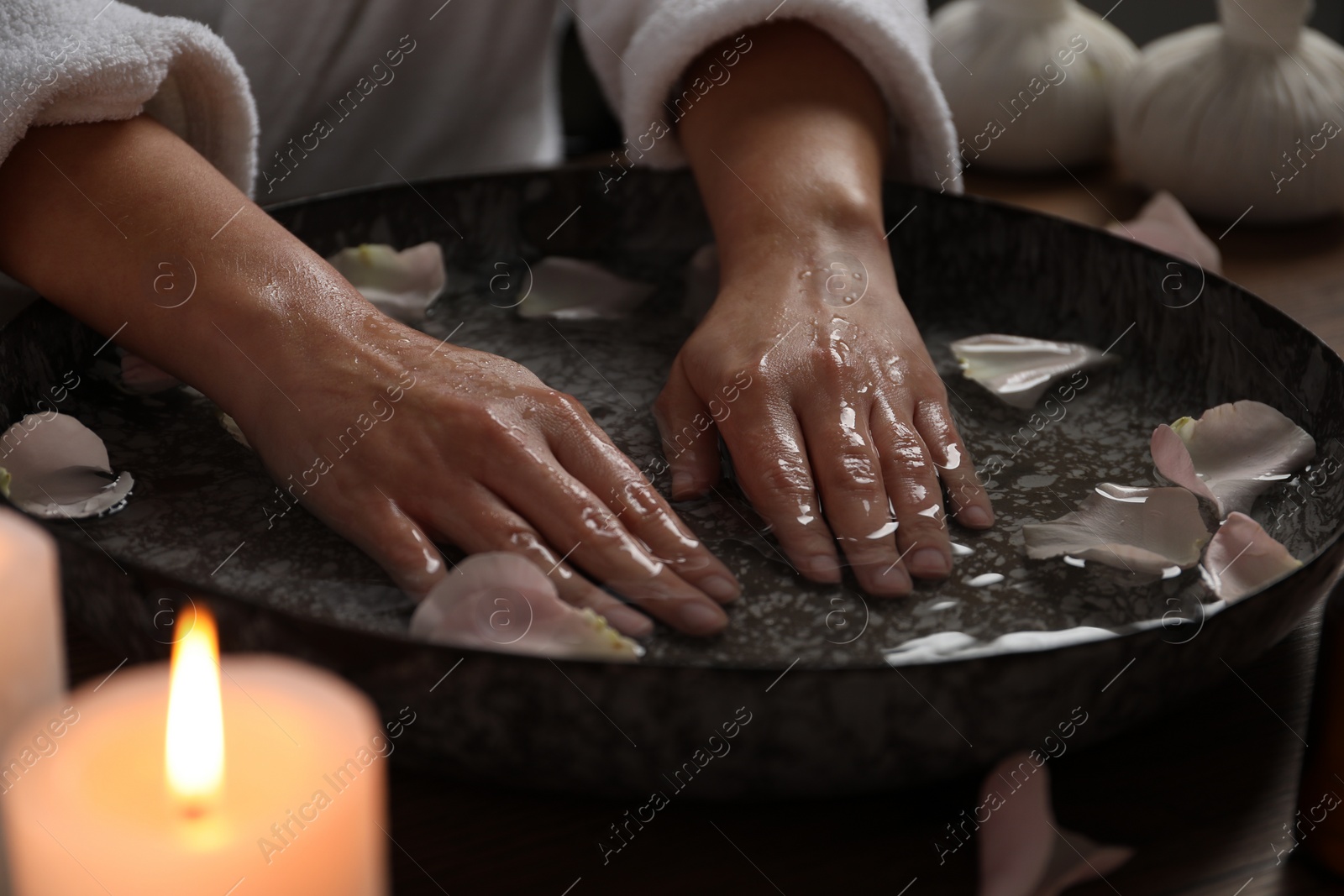 Photo of Woman soaking her hands in bowl of water and flower petals at table, closeup. Spa treatment