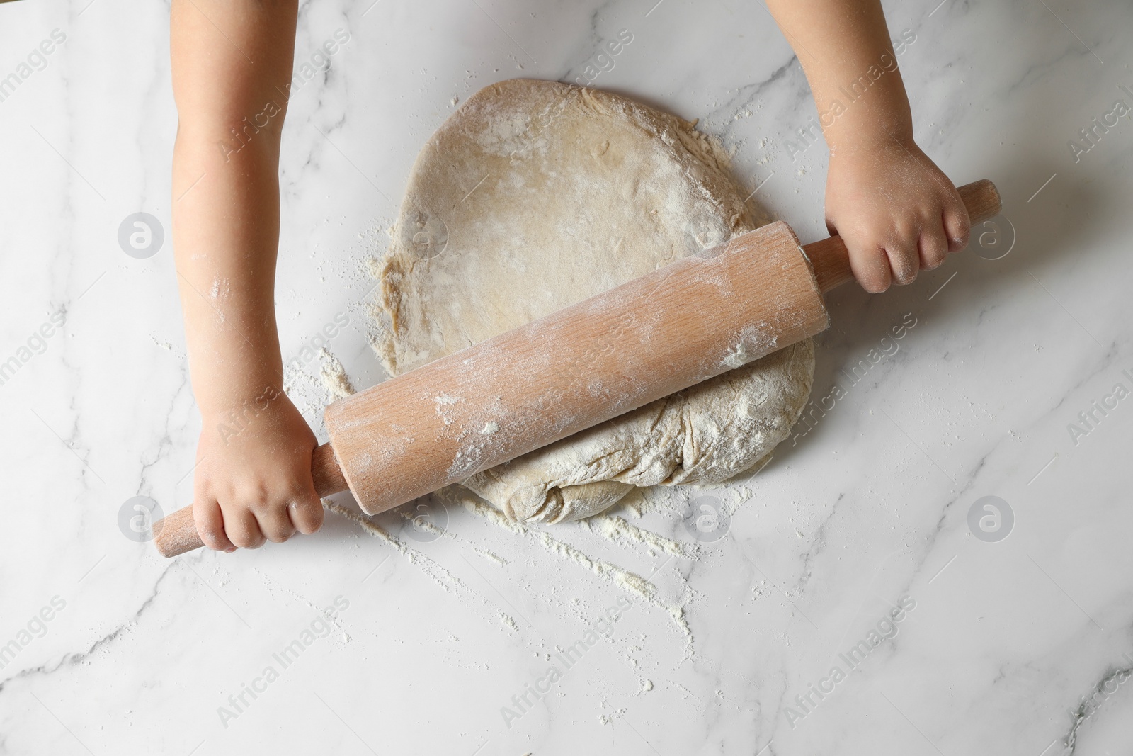 Photo of Little child rolling raw dough at white table, top view