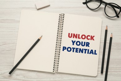 Phrase Unlock Your Potential in notebook, pencils and glasses on white wooden table, flat lay