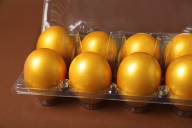 Photo of Shiny golden eggs in plastic box on brown background, closeup