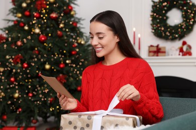 Happy young woman with Christmas gift reading greeting card at home