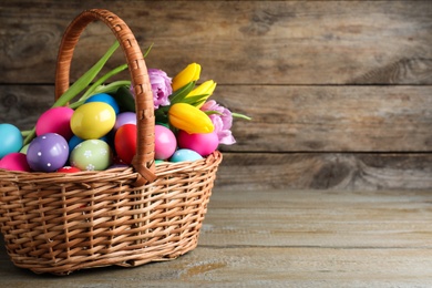 Photo of Colorful Easter eggs and tulips in wicker basket on wooden background. Space for text