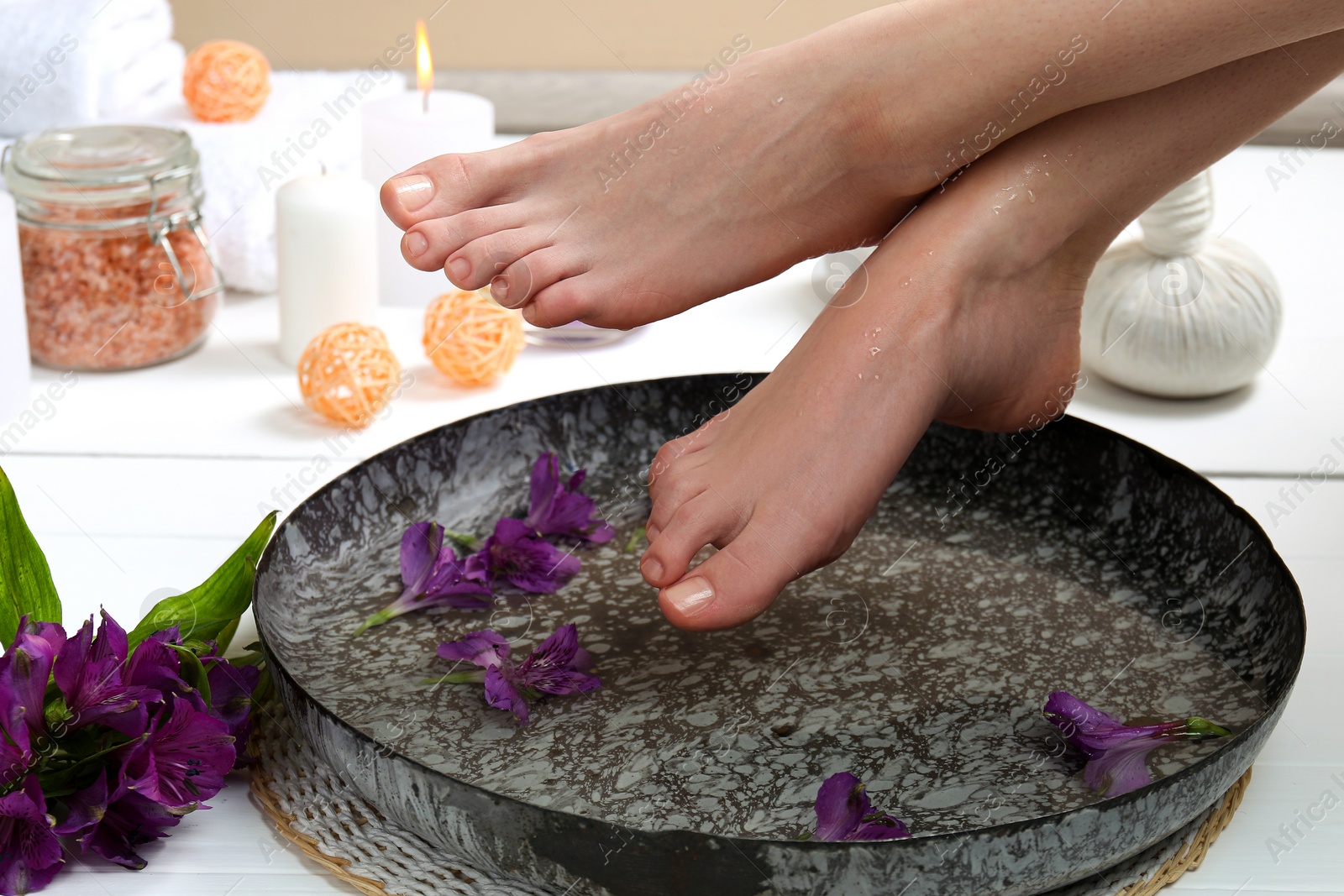 Photo of Woman holding her feet over bowl with water and flowers on white floor, closeup. Spa treatment