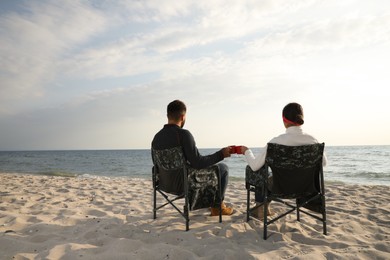 Photo of Couple sitting in camping chairs and clinking mugs on beach, back view