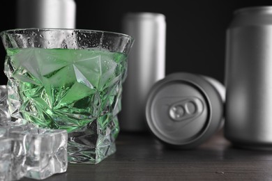 Photo of Energy drink in glass, aluminium cans and ice cubes on wooden table, closeup