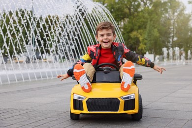 Photo of Cute little boy driving children's car near fountain on city street. Space for text