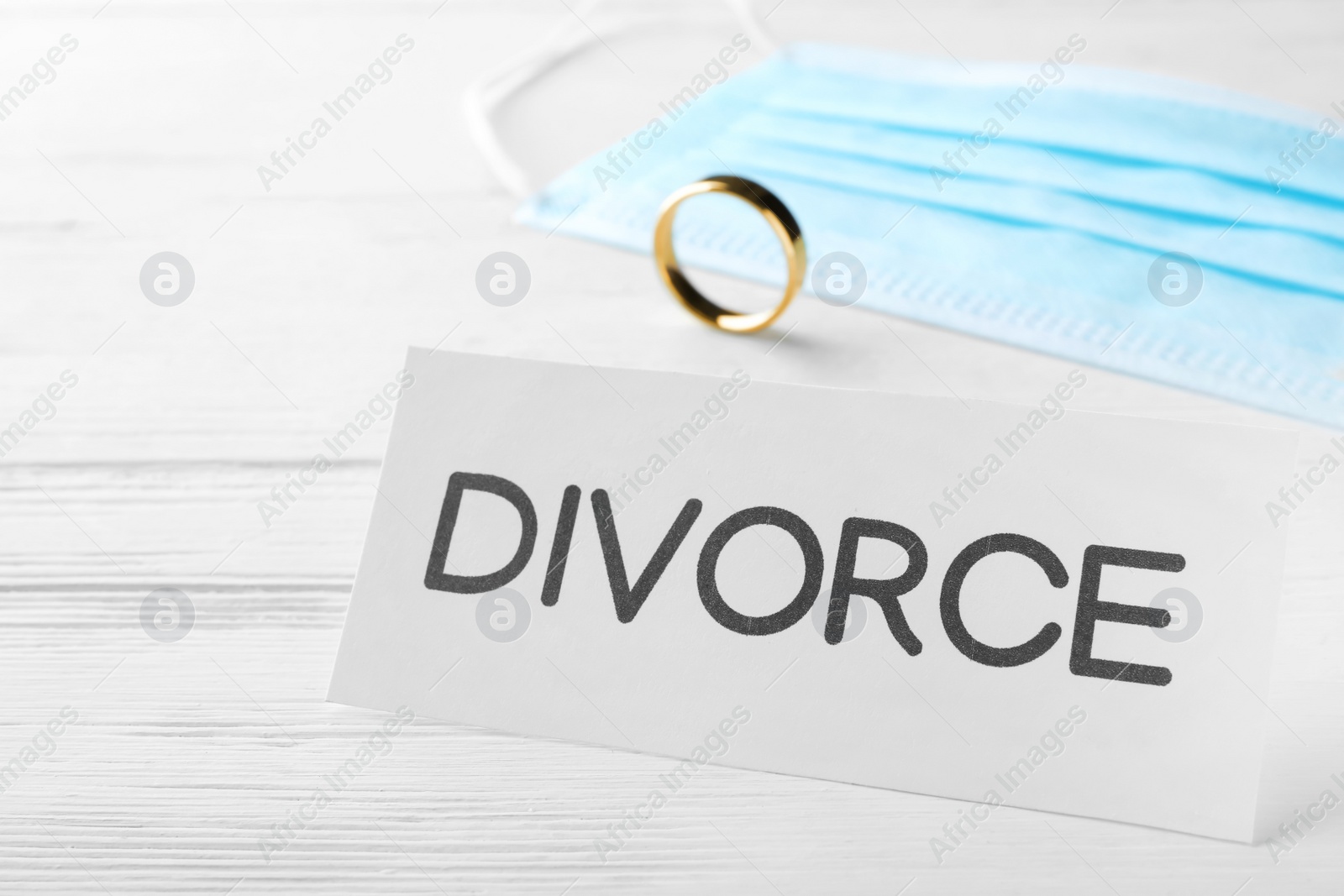 Photo of Card with word Divorce, protective mask and wedding ring on white wooden table, closeup