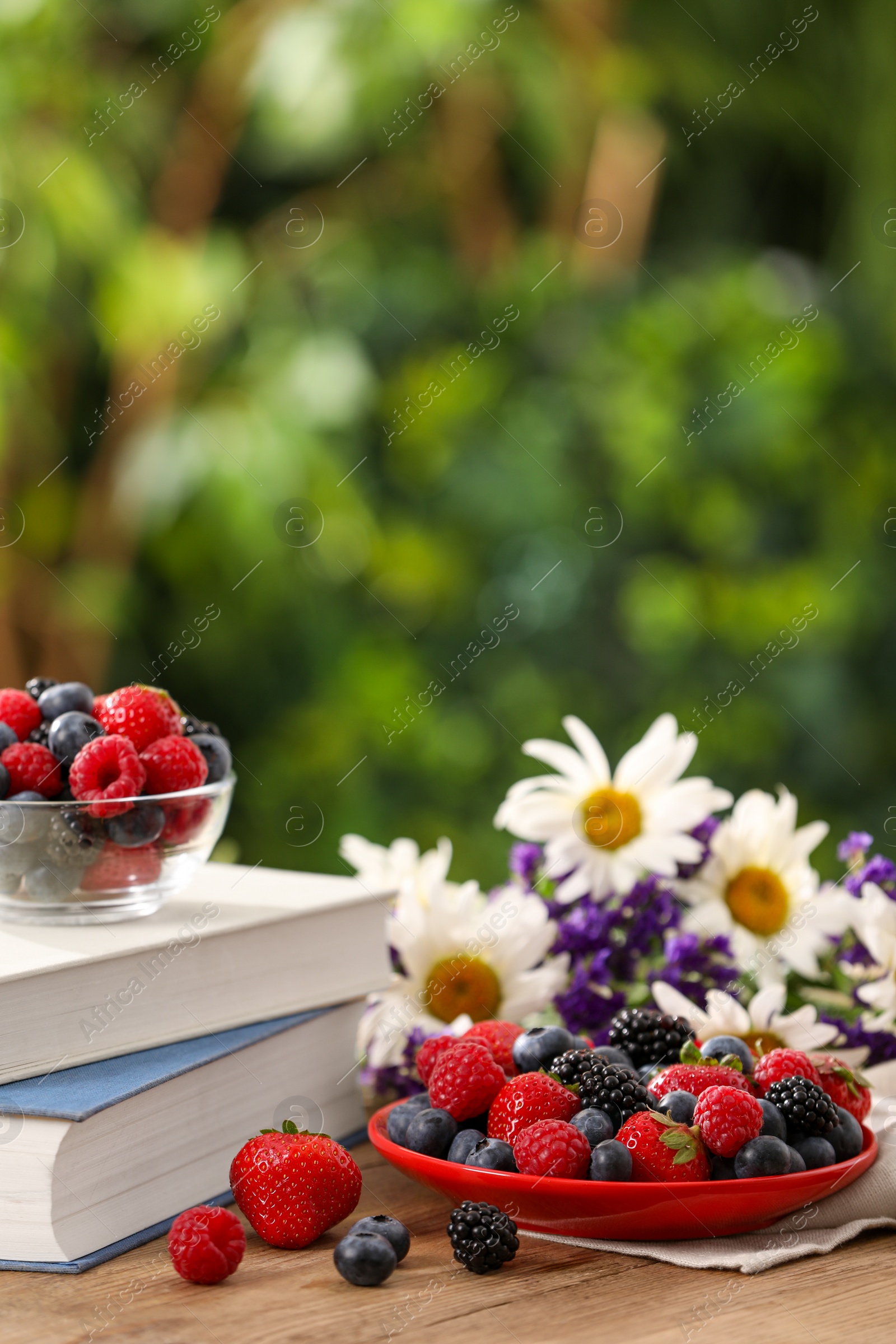 Photo of Different fresh ripe berries, beautiful flowers and books on wooden table outdoors