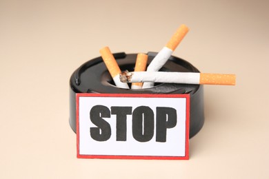 Photo of Card with word Stop, ashtray and cigarette stubs on beige background, closeup. Quitting smoking concept