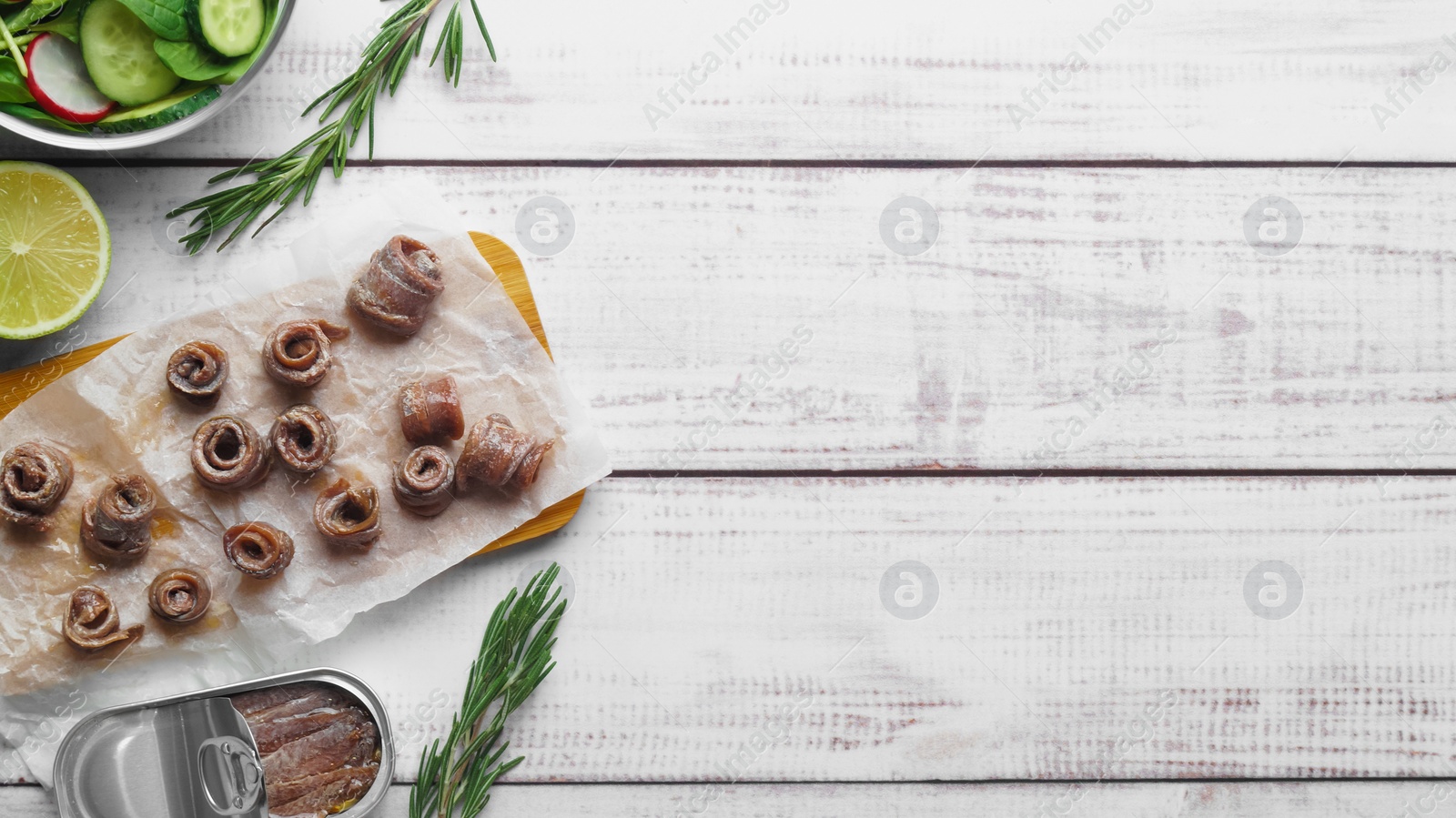 Photo of Canned anchovy fillets, rosemary and lime on white wooden table, flat lay. Space for text