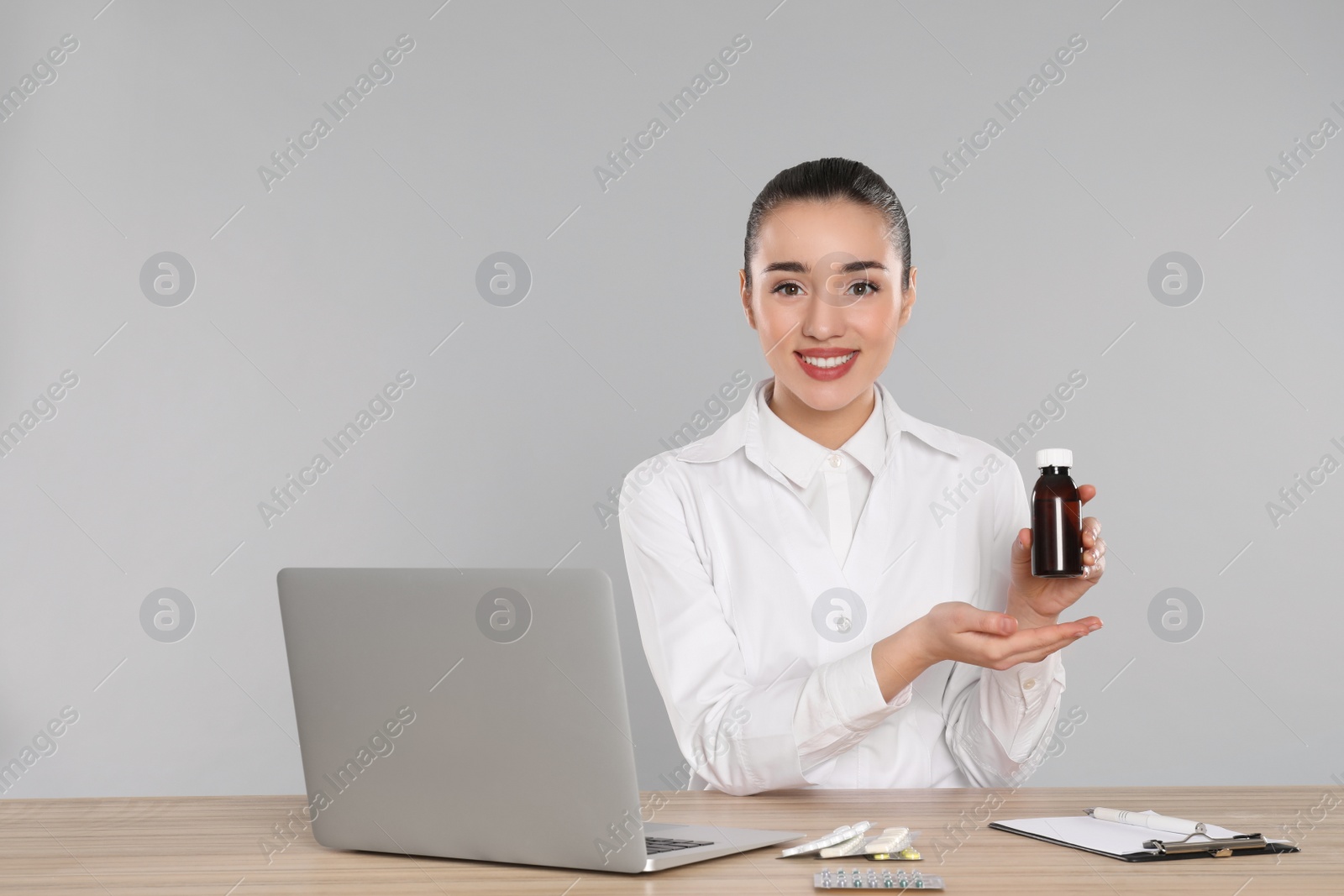 Photo of Professional pharmacist with syrup and laptop at table against light grey background