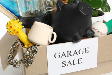 Photo of Different stuff in box with sign Garage Sale on white background, closeup