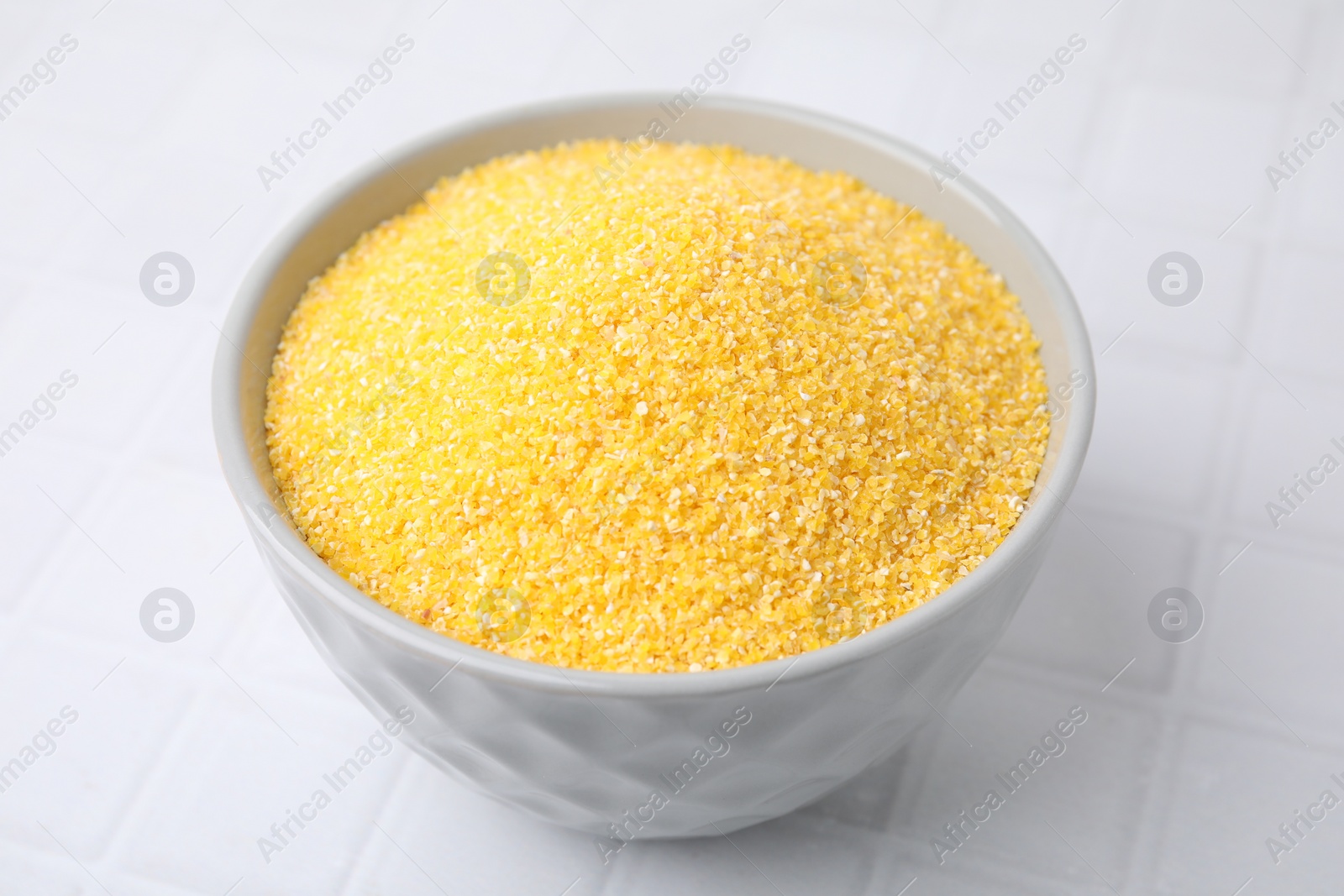 Photo of Raw cornmeal in bowl on white tiled table, closeup