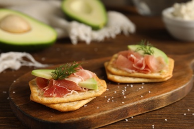 Photo of Delicious crackers with avocado, prosciutto and dill on wooden table, closeup