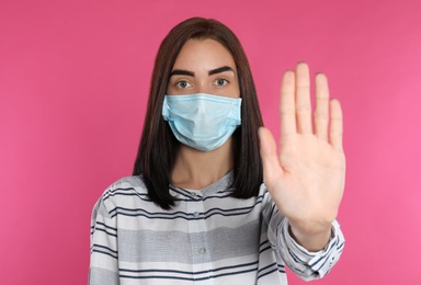Photo of Young woman in protective mask showing stop gesture on pink background. Prevent spreading of coronavirus