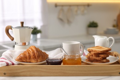 Photo of Breakfast served in kitchen. Tray with toasts, honey, jam, fresh croissant, coffee and pitcher of milk on white table, closeup