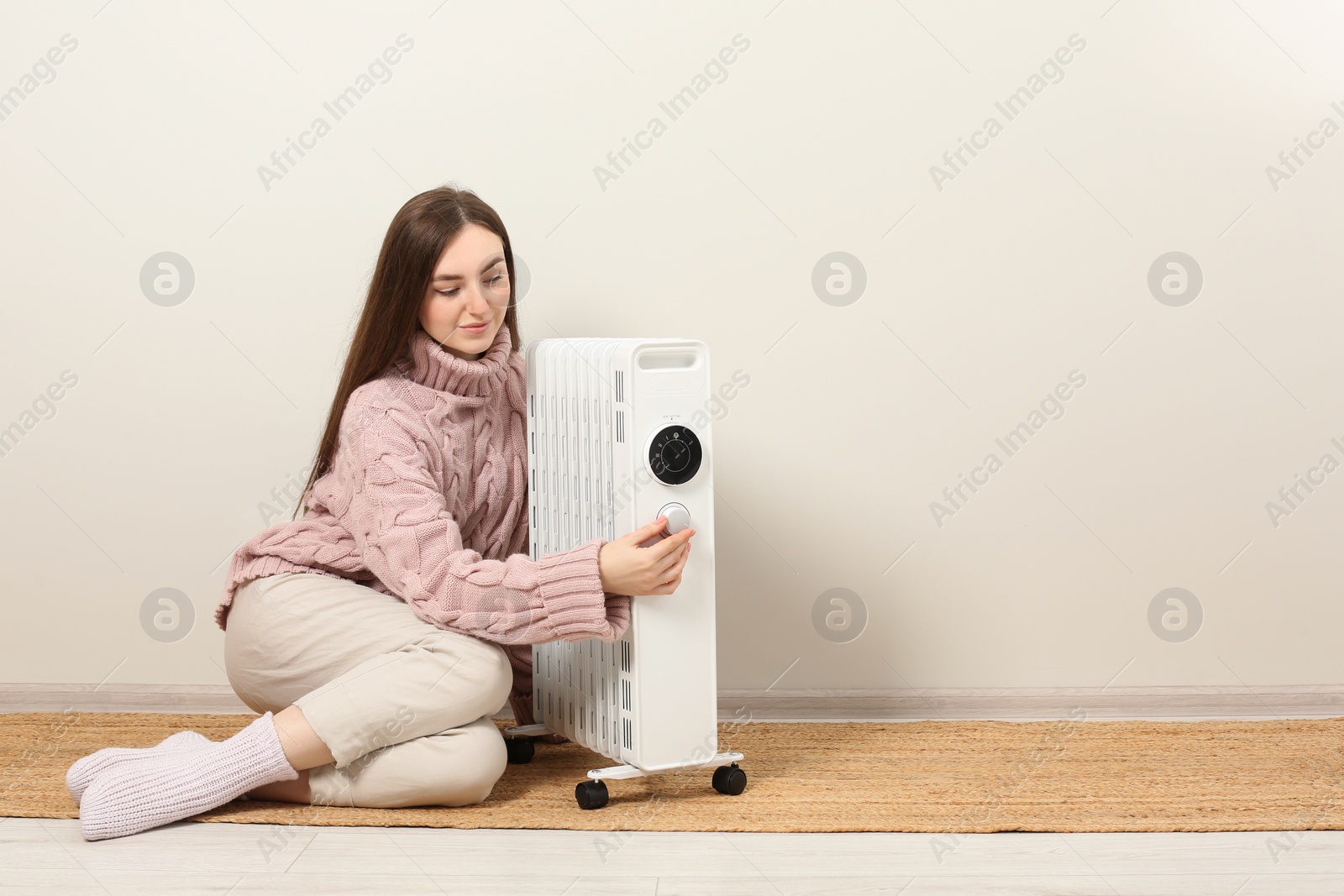 Photo of Young woman adjusting temperature on modern electric heater near beige wall, space for text