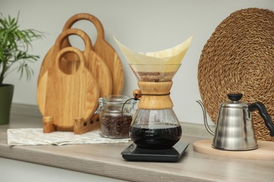 Photo of Making drip coffee. Glass chemex coffeemaker with paper filter, jar of beans and kettle on wooden countertop in kitchen