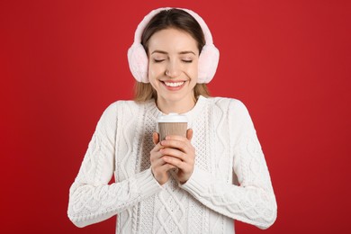 Happy woman with cup of drink wearing warm earmuffs on red background