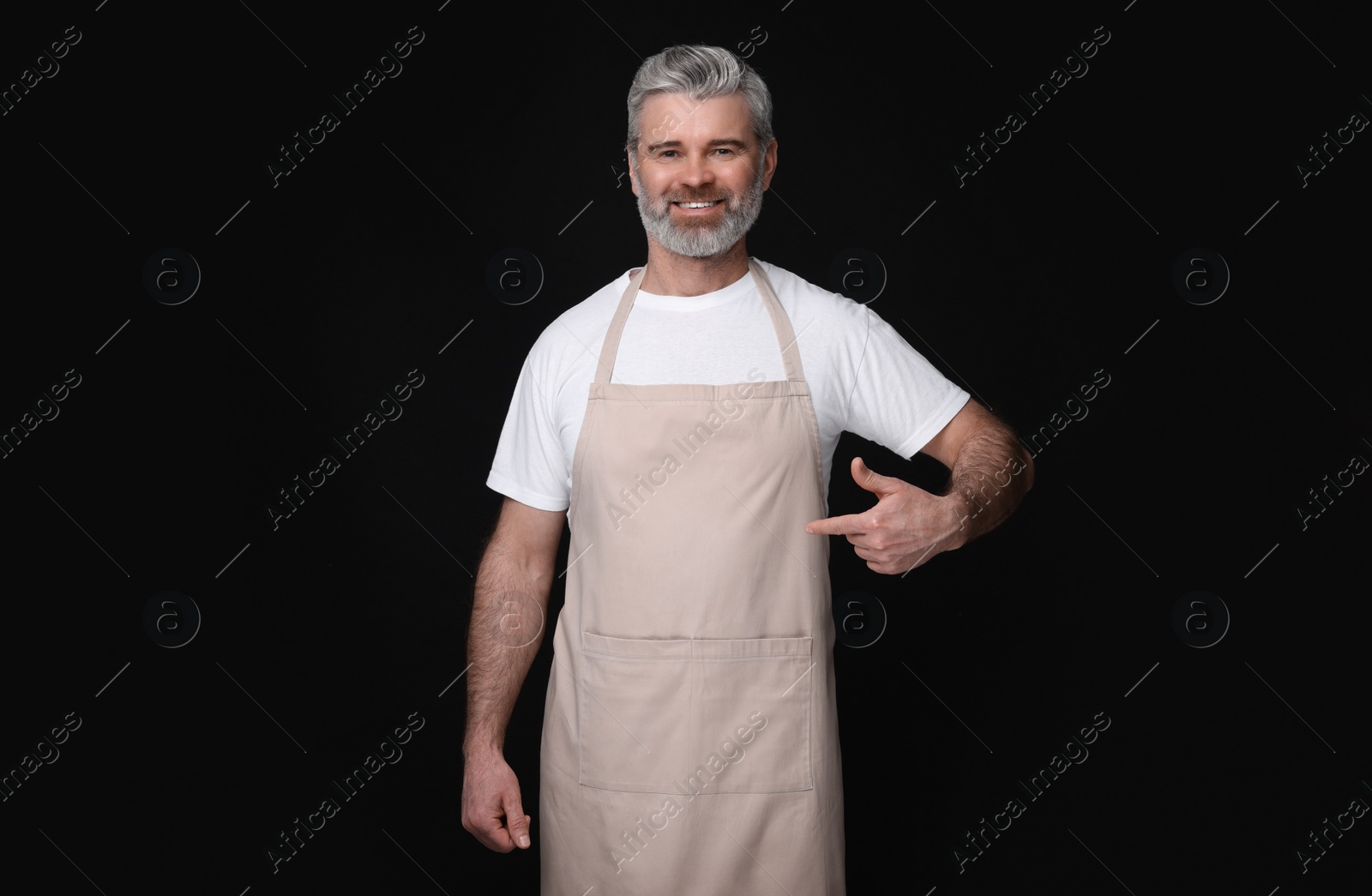 Photo of Happy man pointing at kitchen apron on black background. Mockup for design