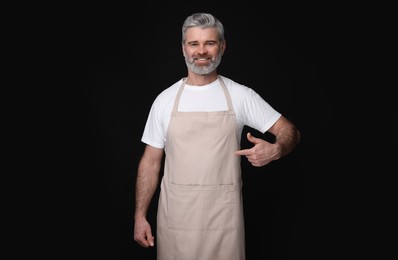 Photo of Happy man pointing at kitchen apron on black background. Mockup for design