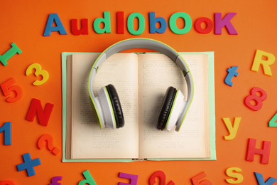 Photo of Word AUDIOBOOK, headphones and open tome on orange background, flat lay