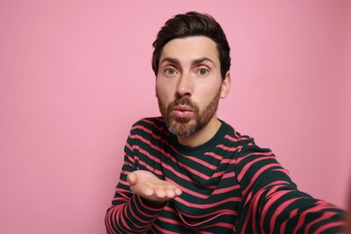 Photo of Handsome man blowing kiss while taking selfie on pink background. Space for text