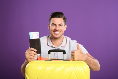 Handsome man with suitcase and ticket in passport for summer trip on purple background. Vacation travel